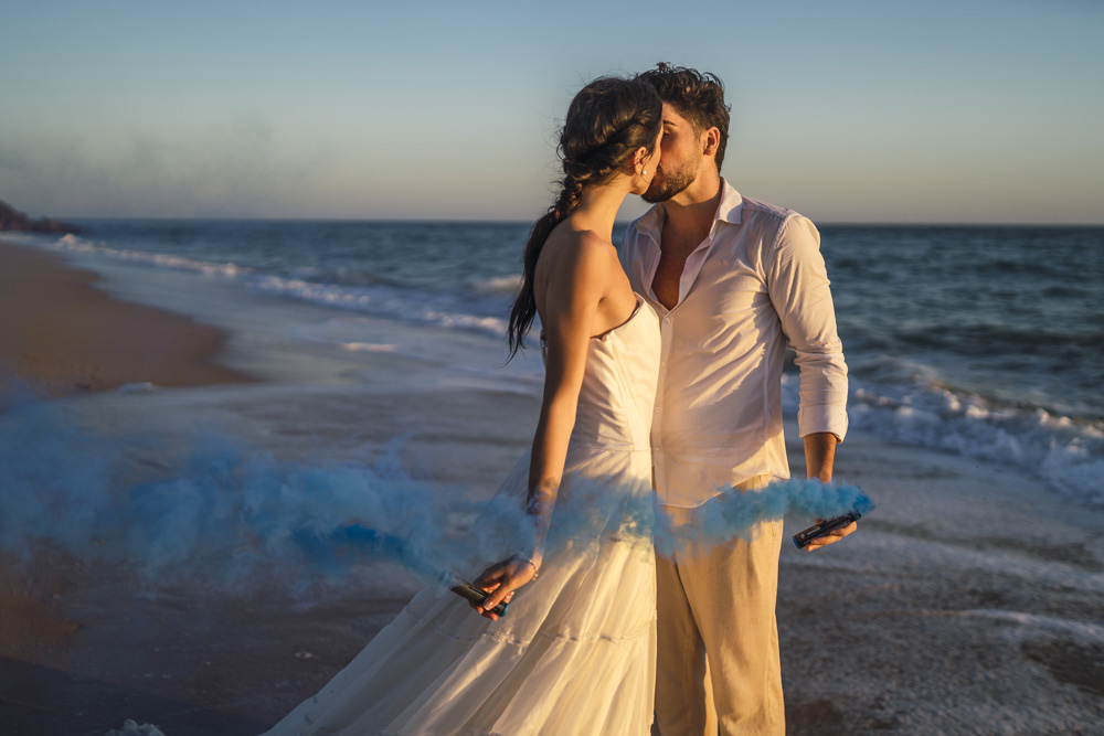 caucasian-loving-couple-holding-blue-colored-smoke-kissing-beach-during-wedding